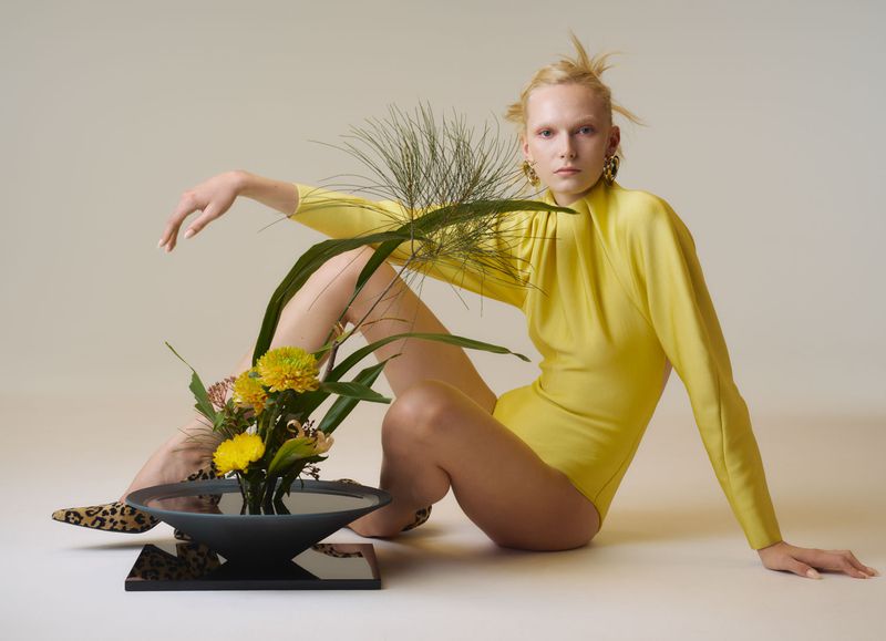 Adult female model in a yellow bodysuit poses with floral arrangement on a light background, creating a fashion and artful visual.