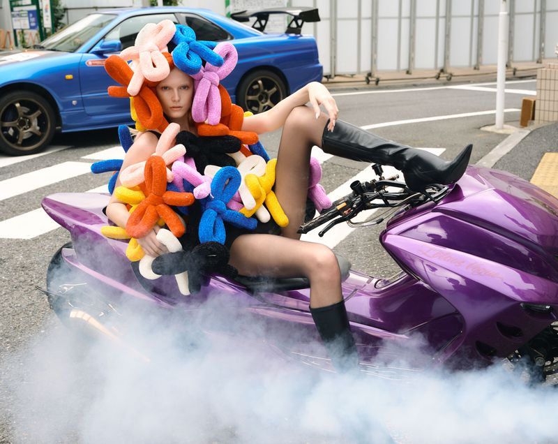 Woman on a purple motorcycle wearing black boots and a costume of colorful stuffed shapes with a blue car in the background.