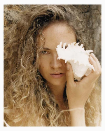 Portrait of a woman holding a conch shell over her eye. The woman has curly hair and is standing outside with a stone background.