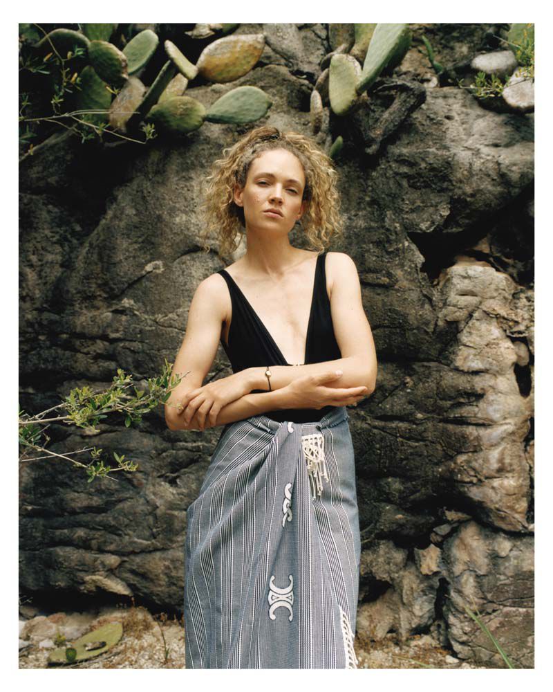 Blonde woman in a black swimsuit and sarong poses outdoors against a rock wall with cacti in a natural environment.