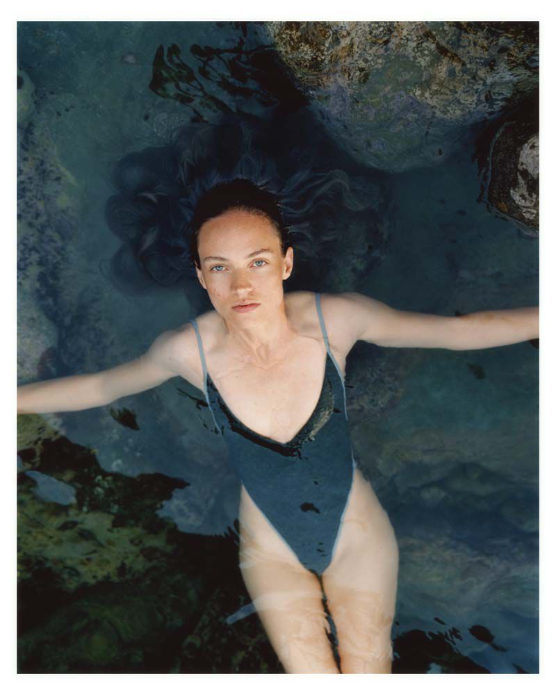 Woman in a blue swimsuit floats in clear water with arms outstretched, surrounded by rocks and dappled light in an outdoor setting.