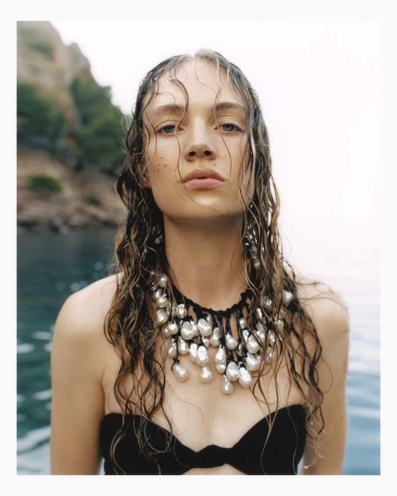 Woman with wet hair, wearing a black top and pearl necklace, stands in water against a backdrop of a green, rocky coastline.