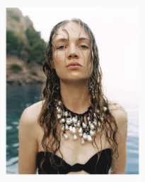 Adrienne Jüliger with wet hair wearing a chunky baroque pearl statement necklace and black swimwear against coastal water.
