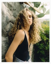 Model Adrienne Jüliger wearing a black swimsuit leaning against a textured rock wall for Grazia UK