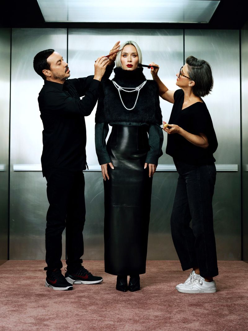 A fashion shoot captures a model in black, with stylists adjusting her jewelry, set against a metallic elevator backdrop and pink carpet.