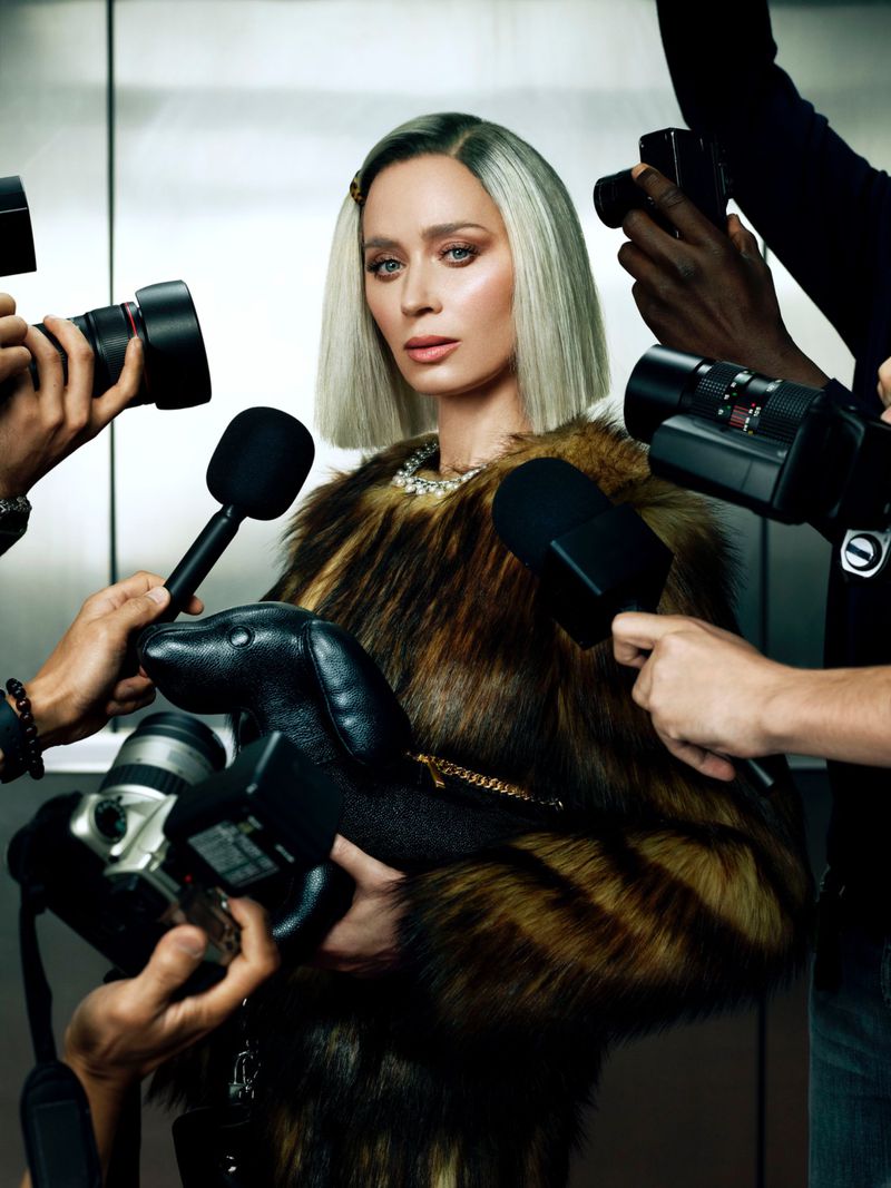 Woman with platinum bob, fur coat, and dog purse is surrounded by photographers in an indoor setting, suggesting a celebrity event.