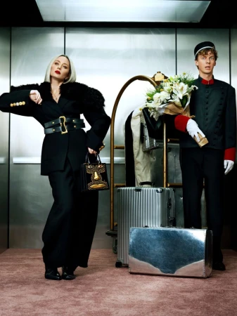 Stylish woman in black suit with bellhop in hotel elevator, carrying luggage and bouquet in fashion editorial style.