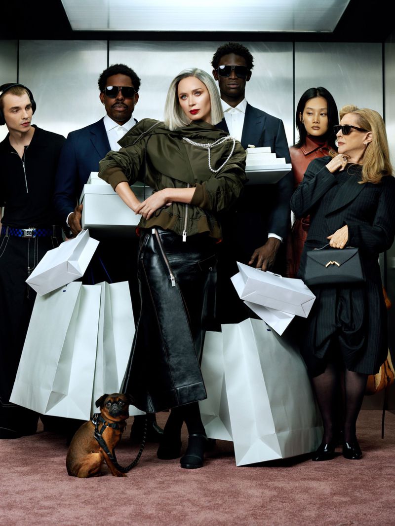Group of fashionably dressed people with shopping bags, including a woman in a green jacket and a small brown dog on a leash.