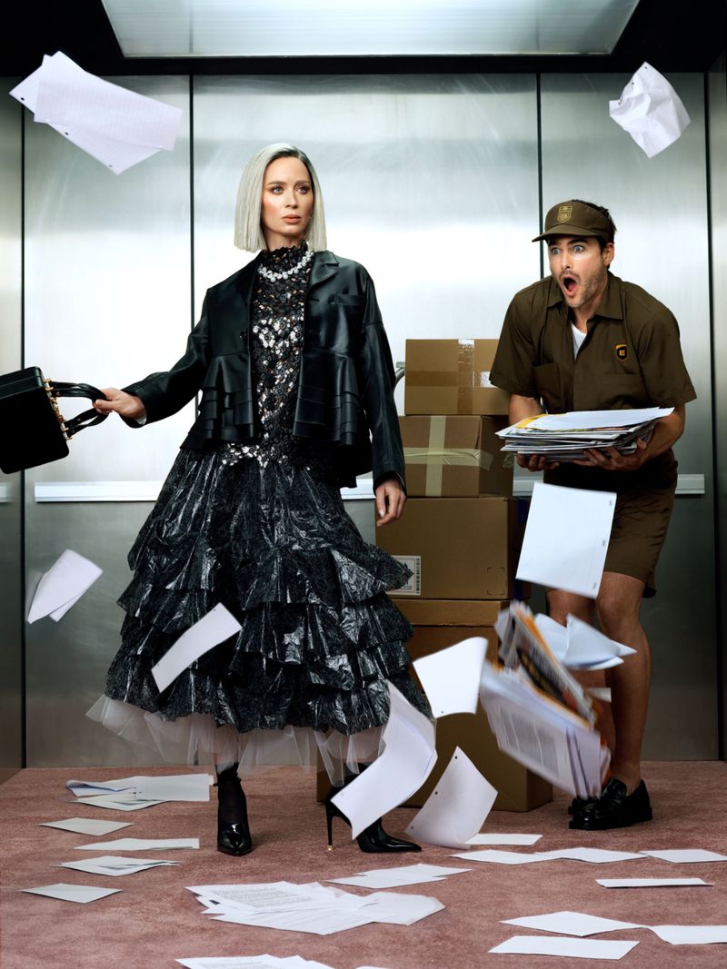 Fashionable woman in a layered black dress drops papers in an elevator, a delivery man in a brown uniform looks on in surprise.