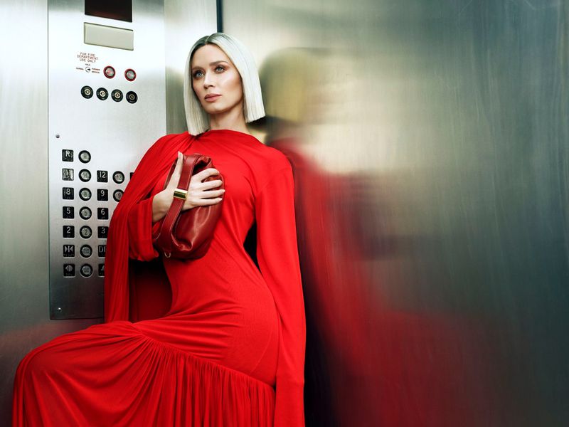 A blonde woman in a long red dress holds a red purse while posing in an elevator with brushed metal walls and fixtures.