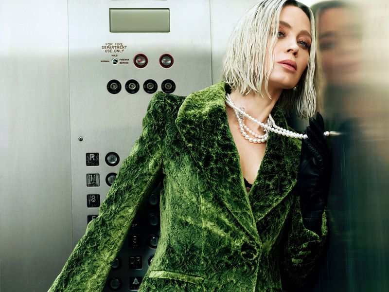 Blonde woman in green textured blazer, black gloves, and pearl necklace poses inside a metal elevator with visible control panel.