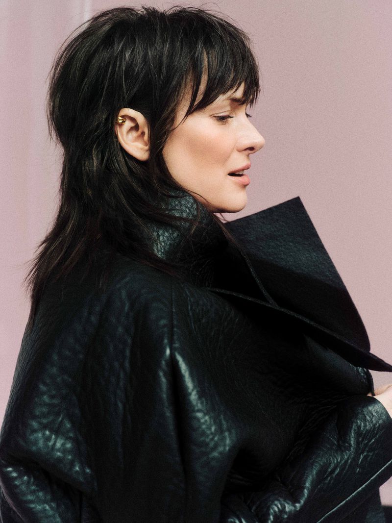 Profile portrait of a woman with dark hair wearing a black leather jacket against a light pink backdrop in a photo studio.