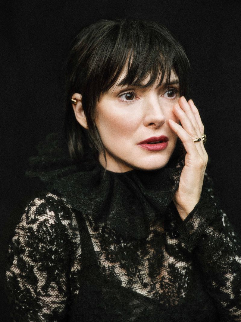 Portrait of a brunette woman in a black lace dress with a ruffled collar against a dark background, with her hand near her face.