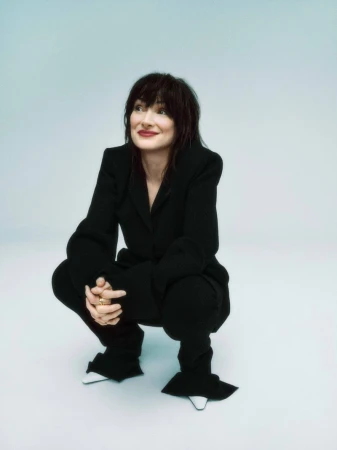 Portrait of a brunette woman in a black pantsuit crouching against a pale blue background, looking up and to the left with a slight smile.