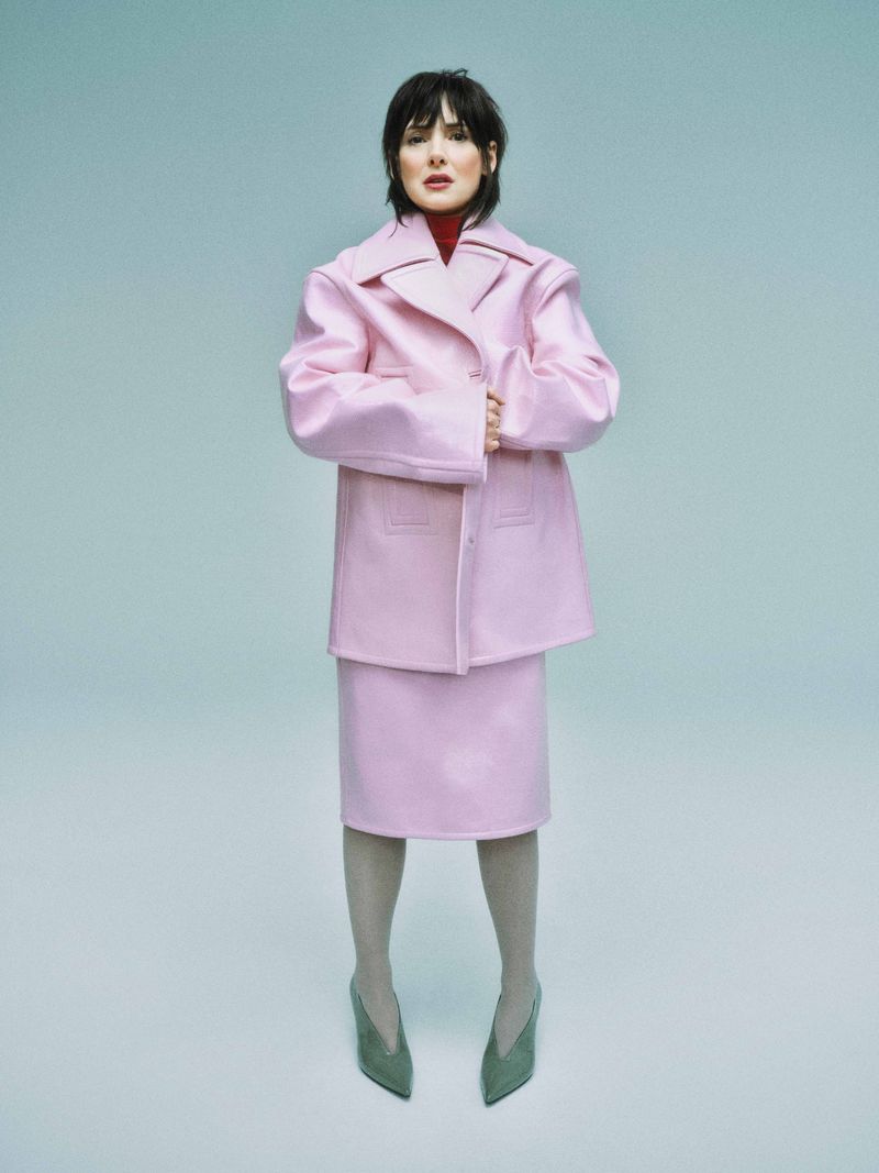 A full shot of a woman in a pink skirt suit standing against a light blue background, wearing gray tights and dark green heels.