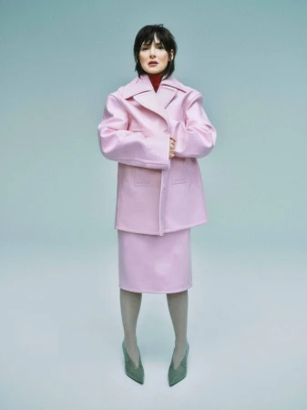 A full shot of a woman in a pink skirt suit standing against a light blue background, wearing gray tights and dark green heels.
