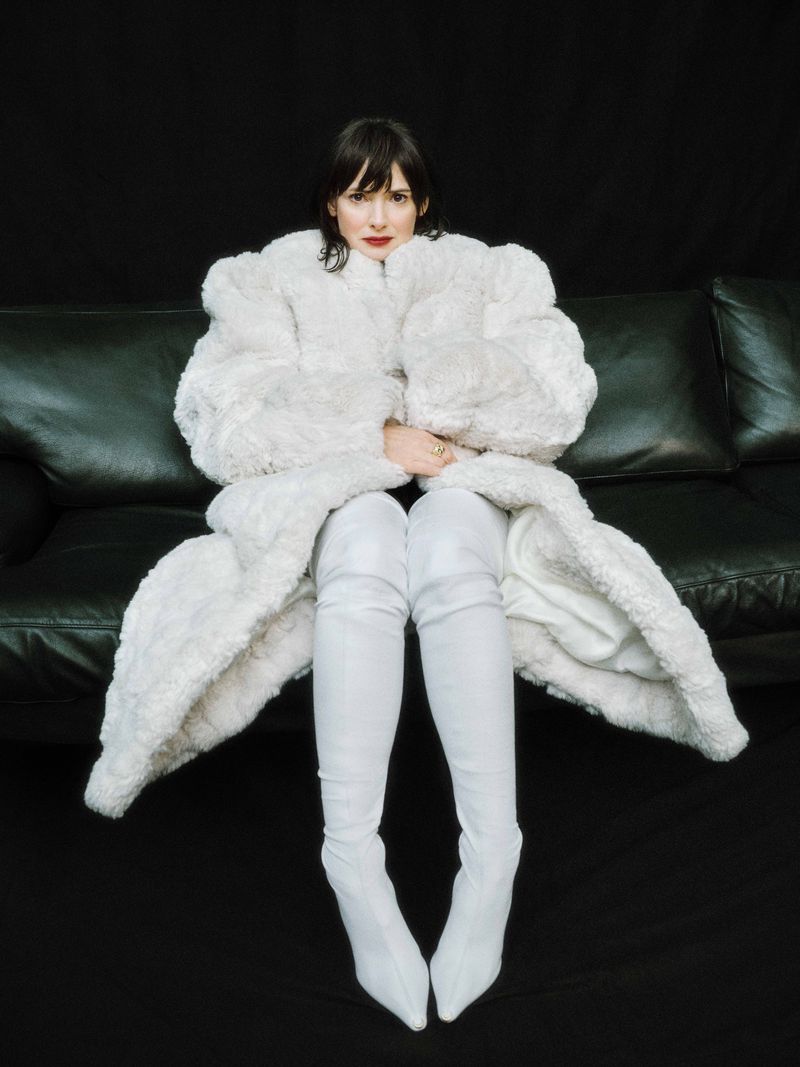 A woman wearing a white furry coat and tall white boots sits on a black leather couch against a black background in a studio shot.