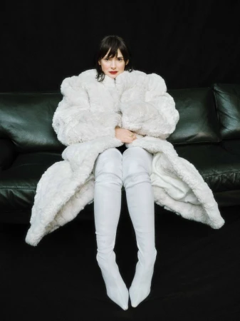A woman wearing a white furry coat and tall white boots sits on a black leather couch against a black background in a studio shot.