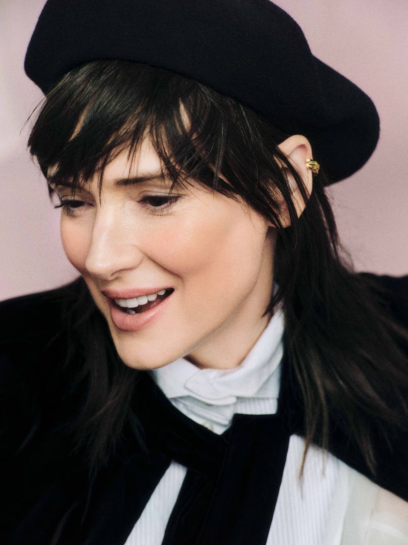Close-up shot of a fair-skinned woman with dark hair, wearing a black hat and white shirt with black bow tie, smiling indoors.