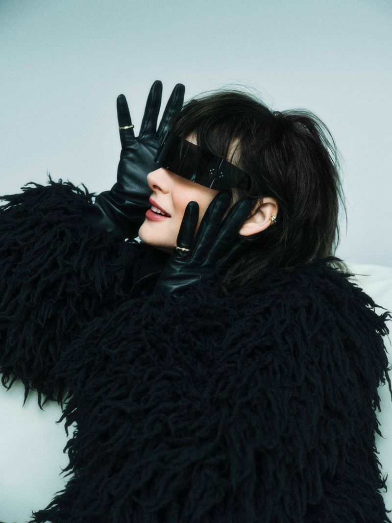 Profile view of a woman in a fur coat, wearing dark glasses and gloves, with her hands up near her face against a plain background.