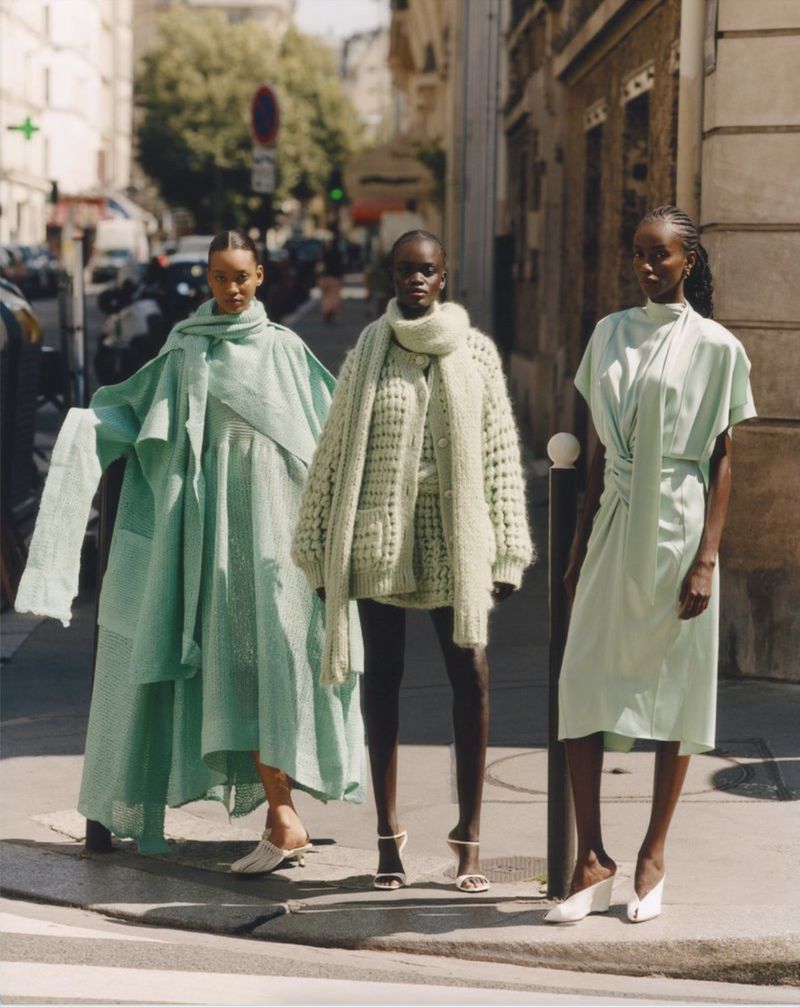Three women stand on a city street wearing pale green outfits. The woman in the center is wearing a heavy, knit sweater.