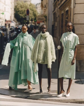 Three women stand on a city street wearing pale green outfits. The woman in the center is wearing a heavy, knit sweater.