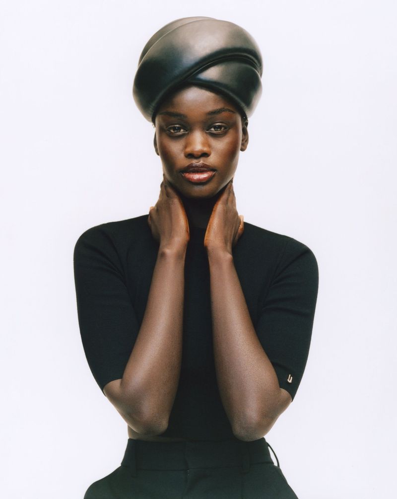Portrait of a Black female model wearing a black turban, turtleneck top, and pants against a white background in a studio setting.