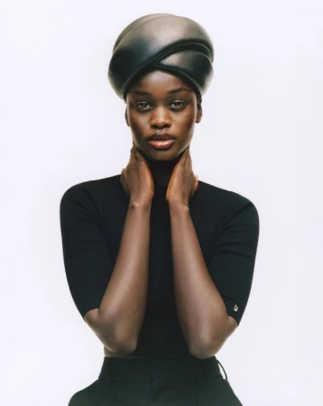 Portrait of a Black female model wearing a black turban, turtleneck top, and pants against a white background in a studio setting.