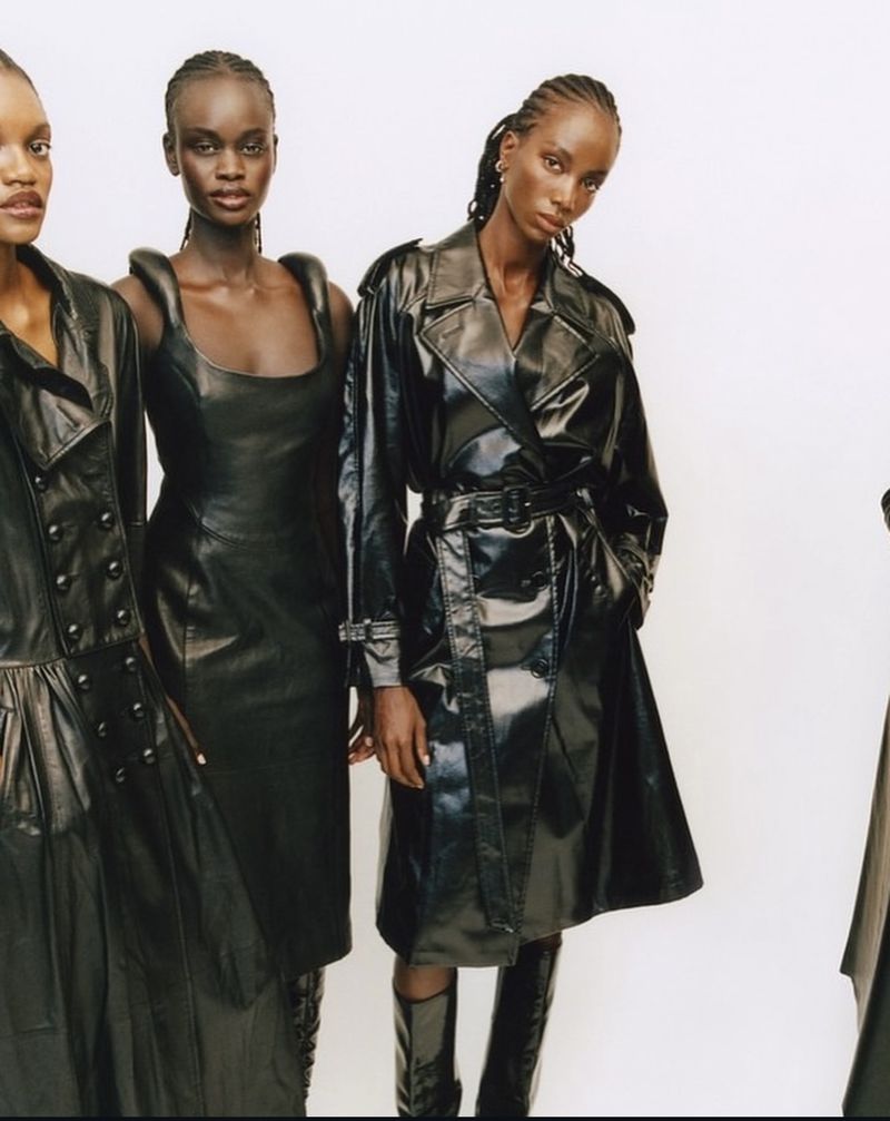 Three African American women in shiny black leather outfits pose in a fashion shot against a white backdrop, showcasing stylish clothing.