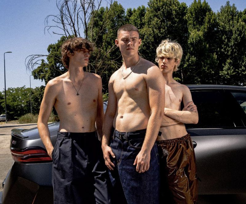 Three shirtless young adult men pose together outdoors in front of a car, showcasing youthful style and summer vibes under bright daylight.