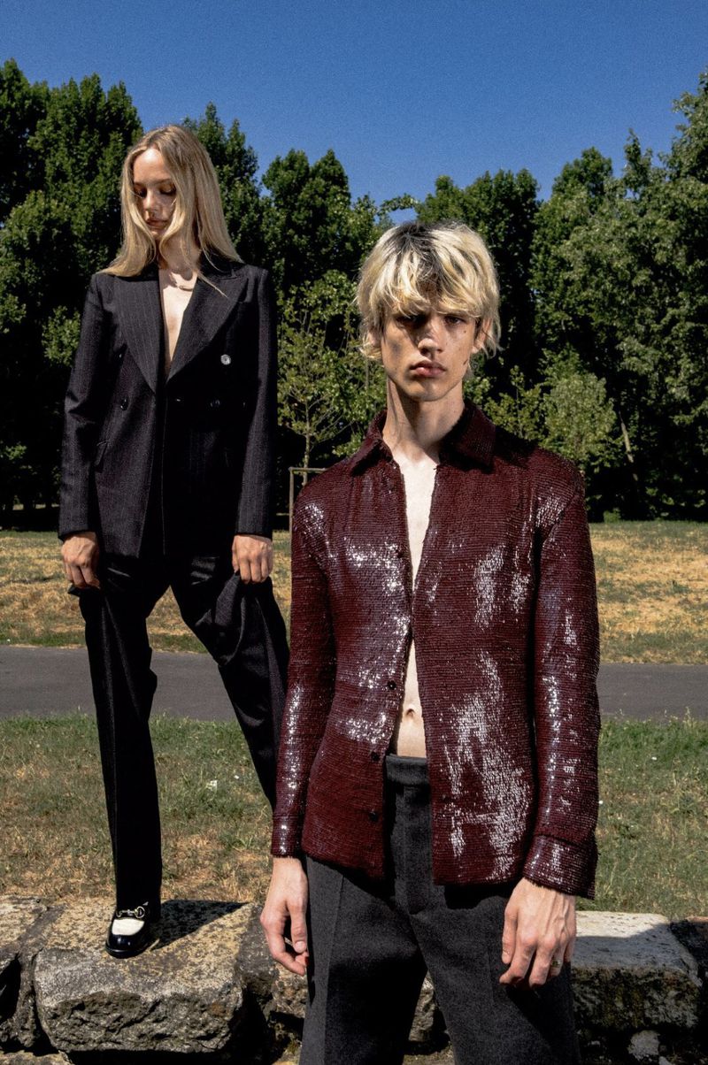 Two fashion models in outdoor setting. Woman wears a black suit, while man sports a burgundy sequined shirt and gray pants in natural light.