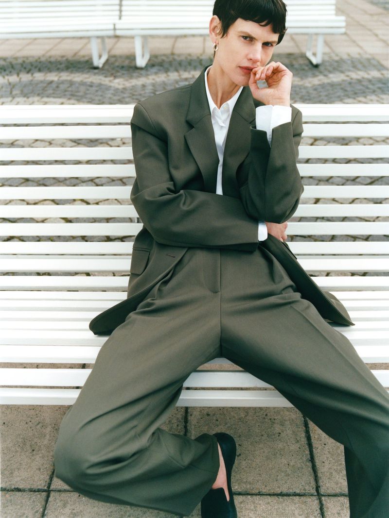 A woman in an olive green suit poses on a white park bench in an eye-level, medium shot, complemented by a neutral background.