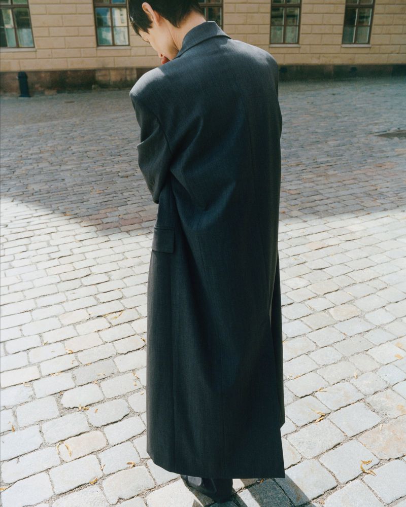A person stands on a cobblestone street wearing a long, dark, oversized coat in front of a light beige stone building with windows.