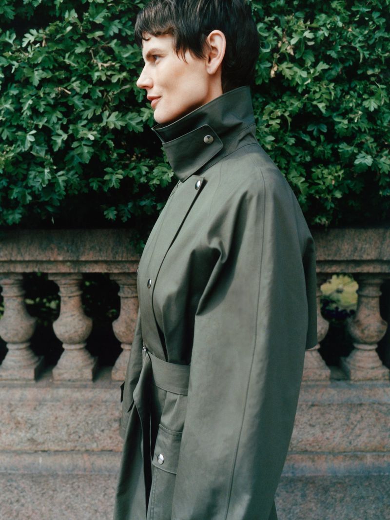 Woman in an olive green trench coat poses outdoors with a greenery background. Fashion shot, portrait photograph. Lifestyle.