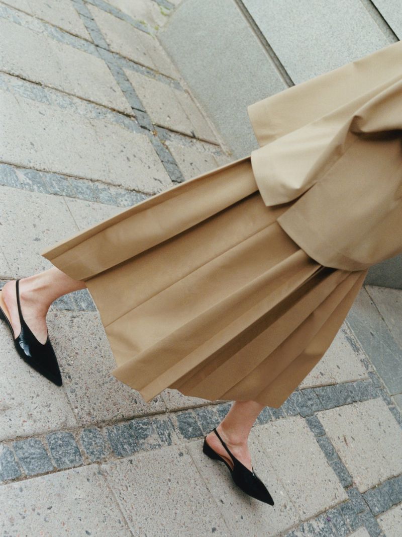 Woman walking wearing a beige pleated skirt and black slingback shoes on stone tiles, fashion editorial, high-angle view.