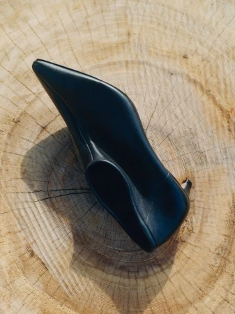 Elevated view of a single black high heel shoe on a textured, light brown tree stump surface, casting a shadow.