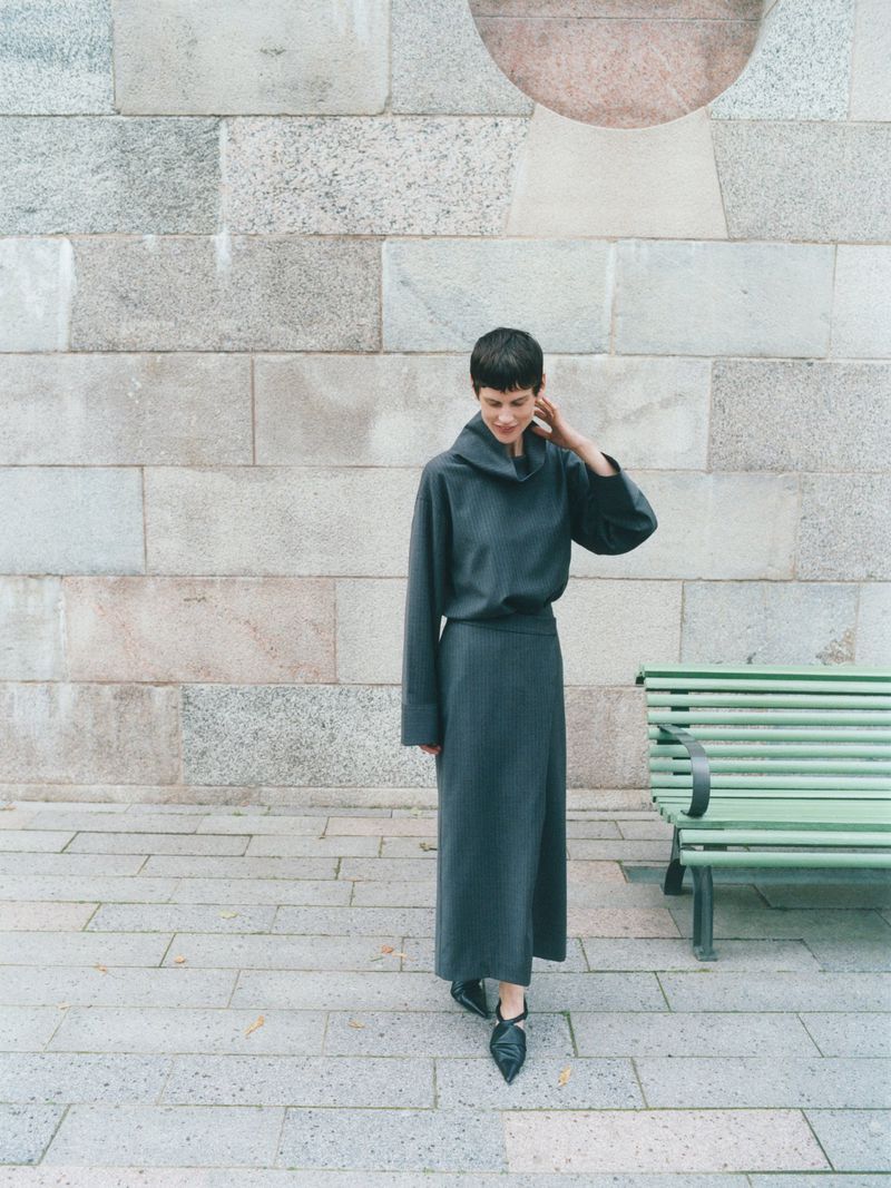 Adult female in a full-length dark-grey dress and black shoes standing outside a building with a bench on a paved walkway.