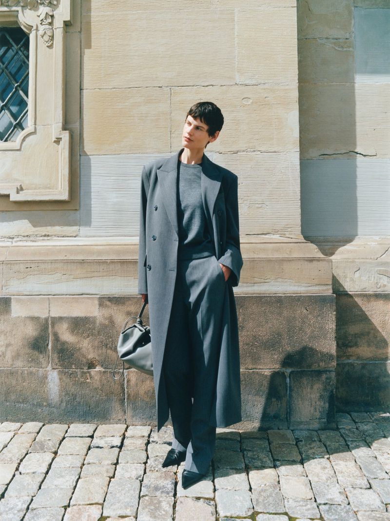 Model standing outdoors wearing a long grey coat and trousers against a stone building wall