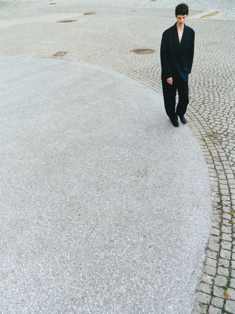 A tall, slender adult walks across a gray-and-white stone plaza. She wears a dark, oversized blazer and slacks. Overhead natural light.