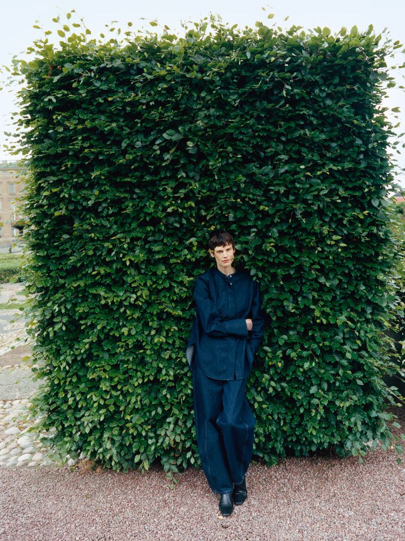 A person in blue clothes poses against a large green hedge with a gravel ground cover in a medium shot in daylight.