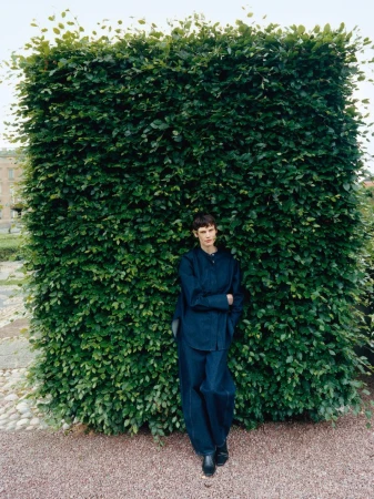 A person in blue clothes poses against a large green hedge with a gravel ground cover in a medium shot in daylight.