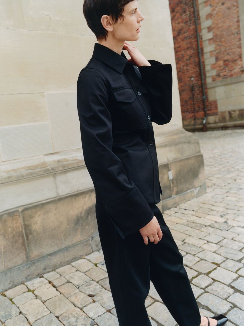 A woman with short dark hair poses outdoors in a black jacket and pants with shoes. The background includes a brick building.