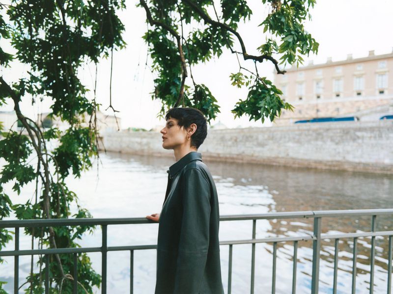 A young adult female in an olive-green jacket standing near a wrought iron railing, with a river and a European-style building in the background.
