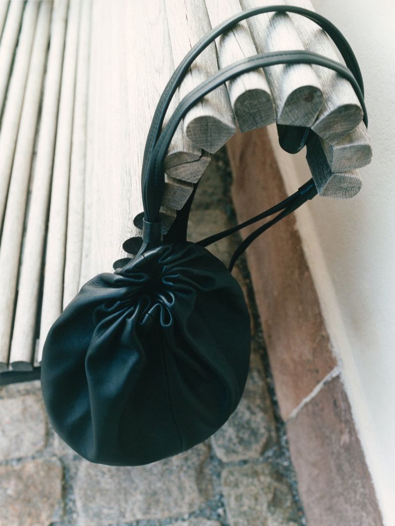 Close-up of a black leather bucket bag with drawstrings and straps, hanging from a light wooden handrail against a stone wall.