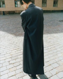 Model seen from behind wearing a dark grey long coat standing on a cobblestone square in front of a stone building exterior