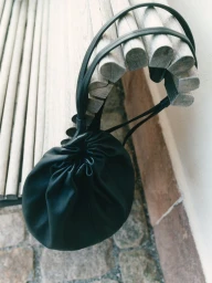 Black leather drawstring pouch handbag hanging from a pale wooden slatted bench outdoors.