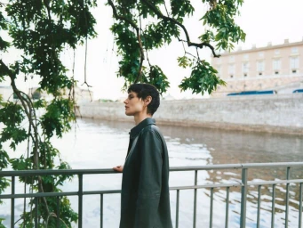 Model with dark hair leaning against a metal railing overlooking a river with buildings and trees in the background