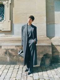 Model standing outdoors wearing a long grey coat and trousers against a stone building wall