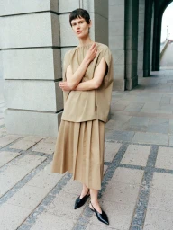Model with short dark hair wearing a beige sleeveless top and pleated skirt outdoors near stone pillars