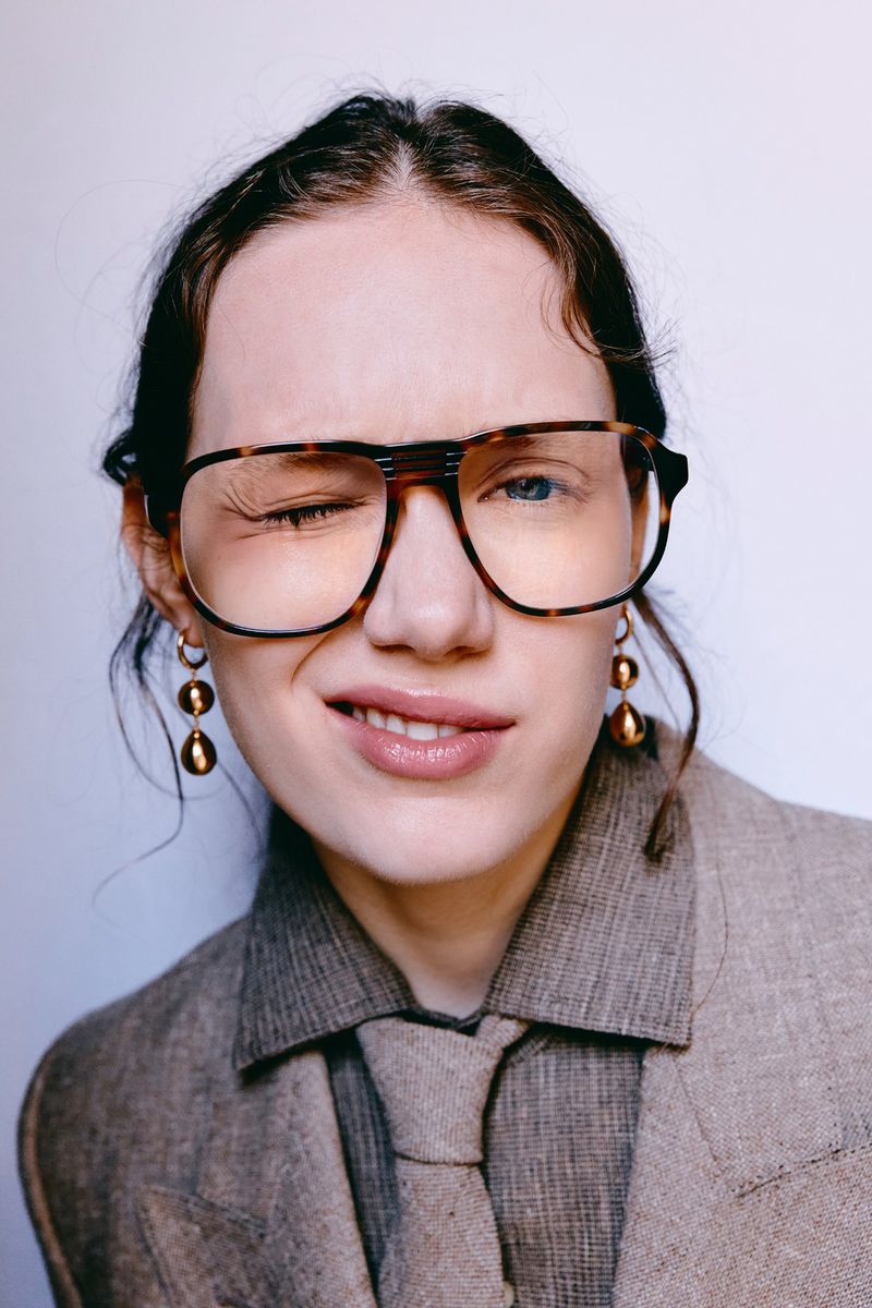 Close-up portrait of a young adult woman winking while wearing large glasses, a gray suit jacket, a gray shirt, and gold earrings.